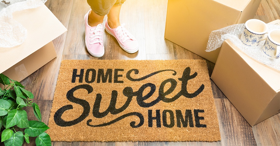 Close up of rug laying on the wood floor that reads “Home Sweet Home” surrounded by moving boxes.