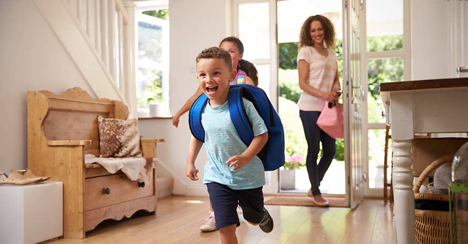 Two young smiling kids wearing backpacks run into foyer of house while mother stands in the doorway smiling.