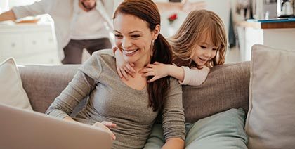 young girl hugging mom as she sits on couch with laptop