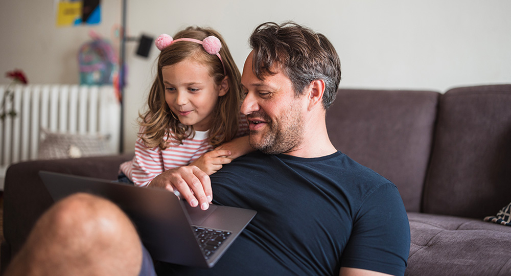 A man and a young child sit together on a couch, smiling as they look at a laptop in their home.
