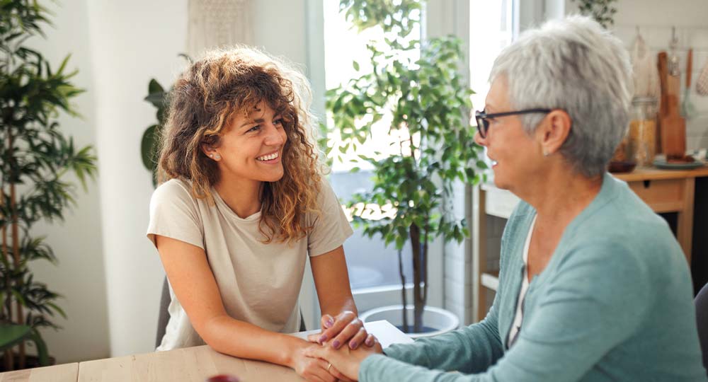 Adult daughter holding hands with her aging mother while discussing extended care planning at home.