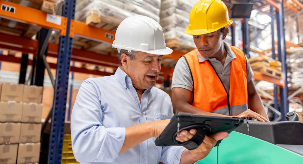 A business owner and a manufacturing worker in safety gear reviewing retirement plan together in a warehouse.