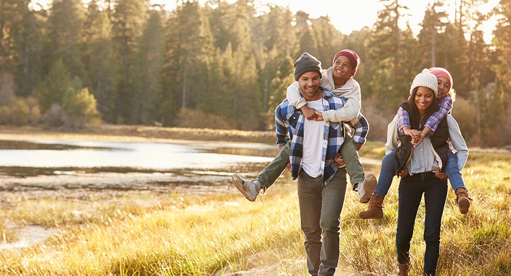 Family walking outdoors. Two parents carry their children on their backs while hiking together on a sunny day.