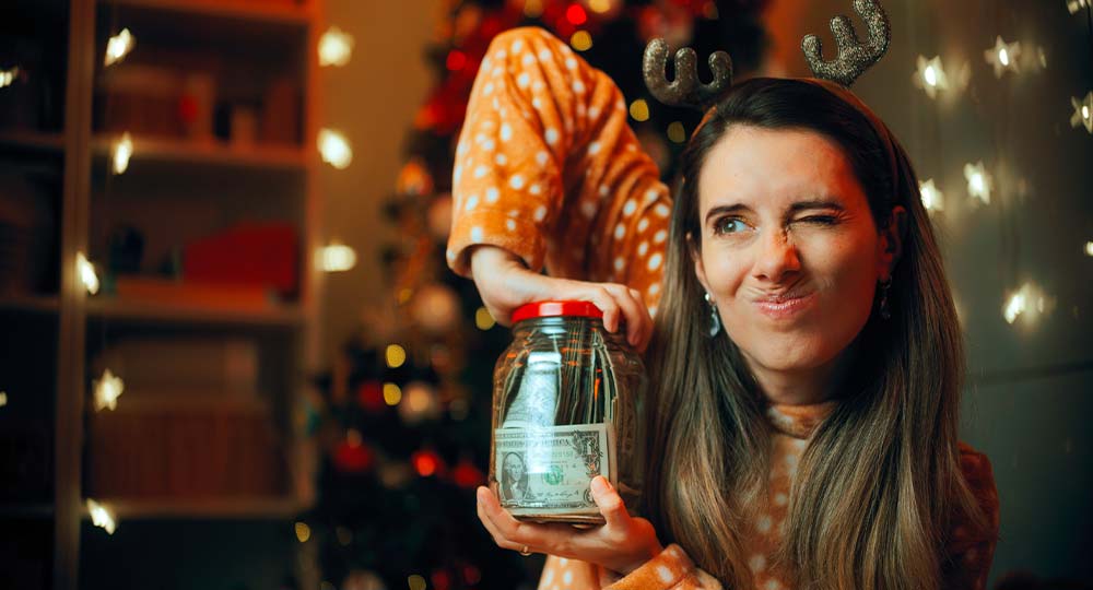 Woman holding a jar of money and making a playful face in front of a Christmas tree, representing holiday budgeting and financial planning.