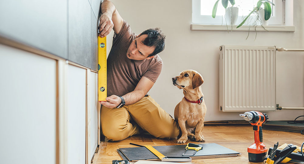 A man kneels on the floor using a level to measure a kitchen cabinet while a dog sits beside him.