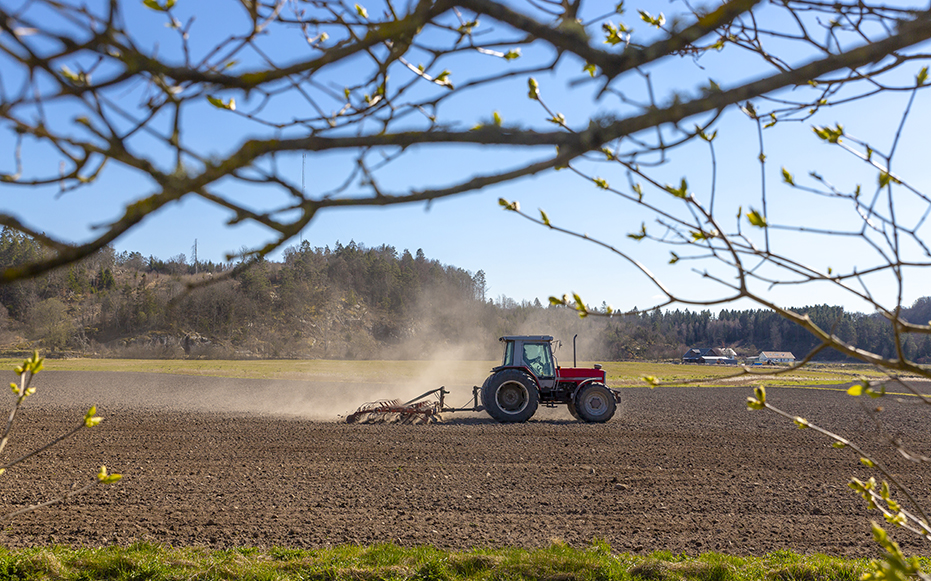 Tractor preparing a farm field in early spring with trees and farmland in the background.