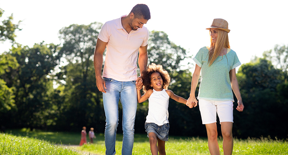 Young parents walking along a green park trail while holding hands with their daughter.